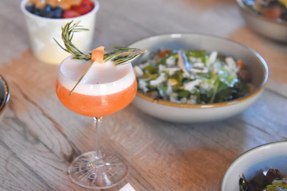 Pink cocktail with a rosemary garnish beside a salad